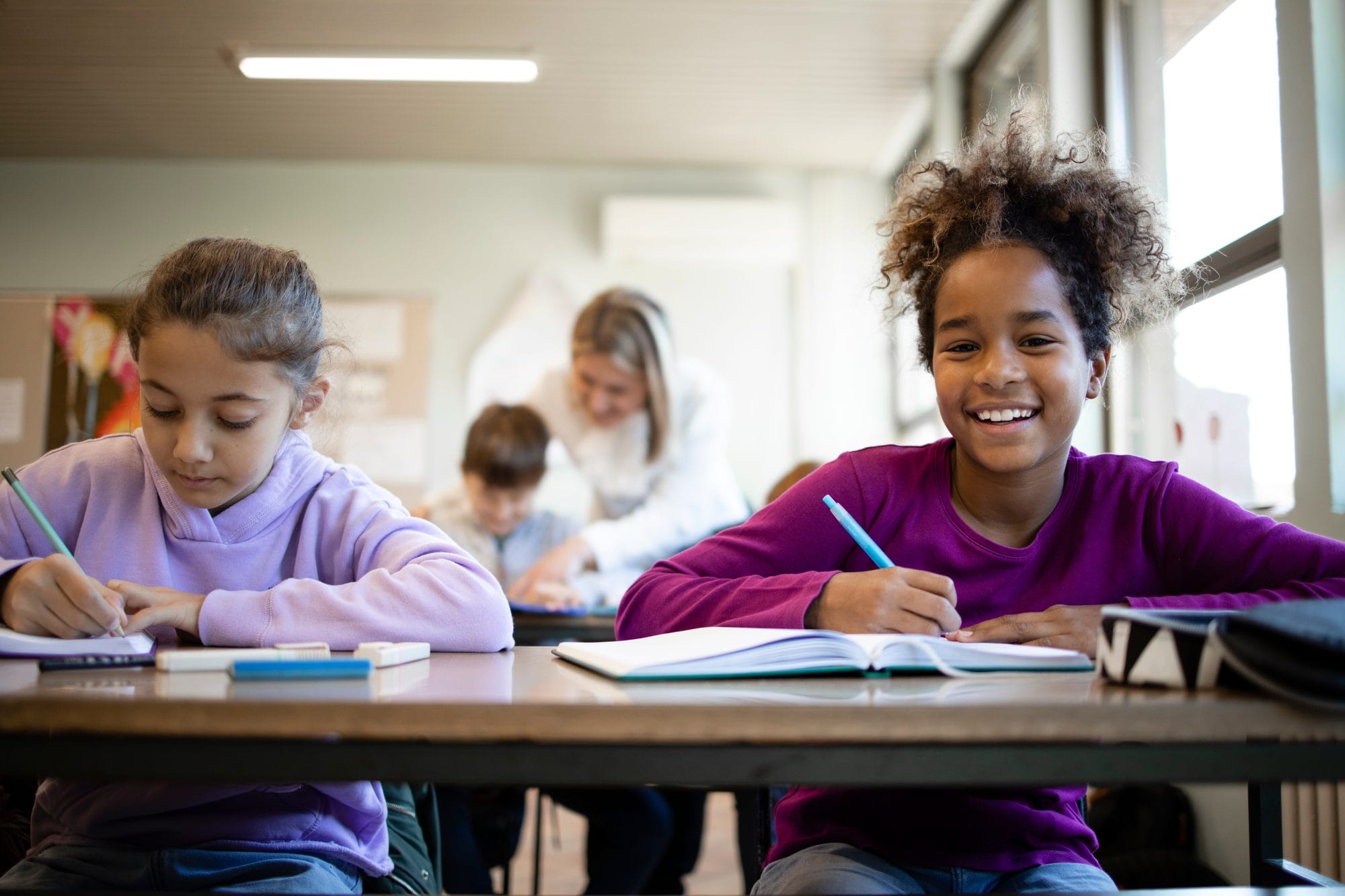 two students in classroom with teacher in the background teaching student